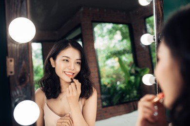beautiful young woman applying glossy lipstick on lips in front of a make up mirror at home