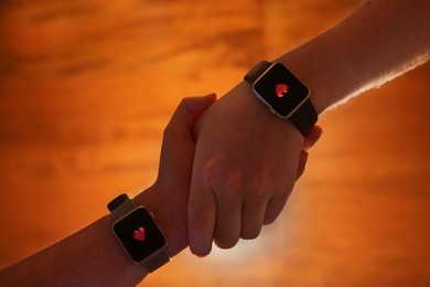 a couple hold hands whilst with hearts on their smart watches against a wooden background.