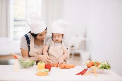 cute little girl and her mom in chef's hats are cutting vegetables cooking a salad and smiling