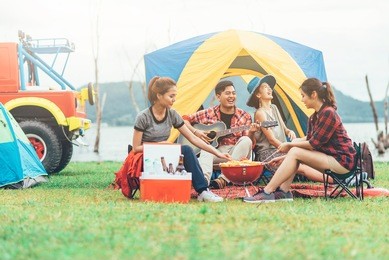 group of asian friends having fun eating barbecue outdoor while camping and play guitar.