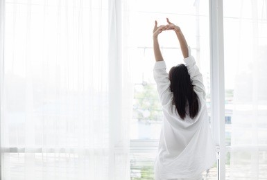 asian woman waking up in her bed fully rested and open the curtains in the morning to get fresh air.