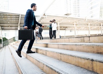 lifestyle of young asian businessman holding black business briefcase walking up the stair with blurred business people background outside office in the city after workout. business concept copy space