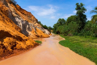 big yellow stream called fairy stream is between special rock wall and green land under blue sky in mui ne , vietnam.