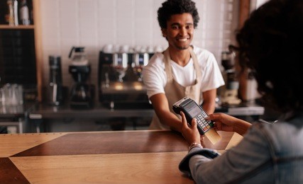 young woman paying by credit card at cafe. woman entering security pin in credit card reader with male barista standing behind checkout counter at coffee shop.