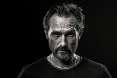 black and white photo of mid aged man showing severe emotion. beardy male in black t-shirt on dark background with serious look.