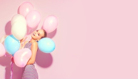 beauty girl with colorful air balloons laughing over pink background. beautiful happy young woman on birthday holiday party. joyful model having fun, playing and celebrating with pastel color balloon