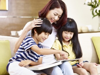 young asian mother and children sitting on couch at home reading a book together, happy and smiling.