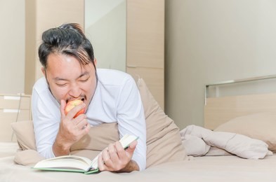 asian man take a relaxing vacation by enjoy to reading book and eating apple in the room.life in room concept