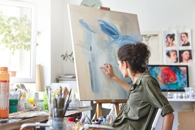 art, creativity, hobby, job and creative occupation concept. rear view of busy female artist sitting on chair in front of easel, painting with fingers, using white and blue oil or acrylic paint