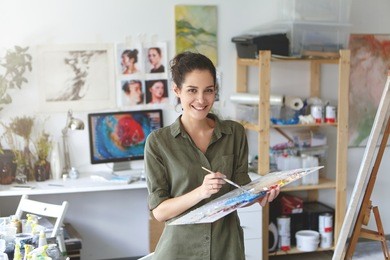 indoor shot of beautiful brunette female painter wearing shirt, holding paint brush in hands standing near easel, creating masterpiece, smiling pleasantly into camera while being glad to paint