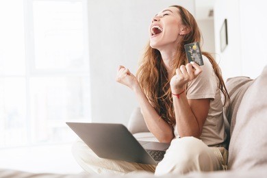 happy young woman with credit card and laptop sitting on sofa at home