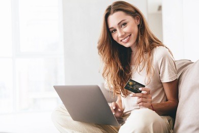 beautiful carefree woman in casual showing credit card to camera while working with laptop on sofa