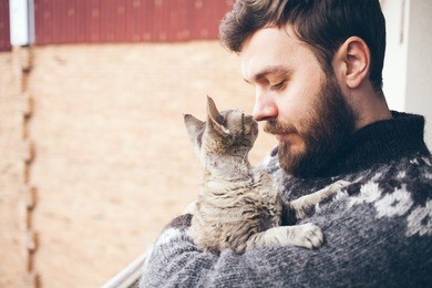close-up of a young man who is standing on a balcony with his cat. home pets. beard man in icelandic sweater is holding and hugging his cute curious devon. copy-space area. home pets, lifestyle.