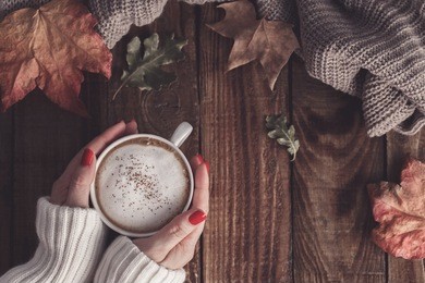 female hands holding hot coffee cappuccino, blanket and autumn leaves on old wooden background. vintage toning. autumn relax concept