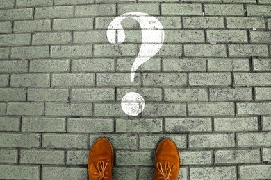 person's feet in suede shoes is standing at tile pavement crossroad with white question mark print pointing. make decision which way to go and what to do. top view. minimalist image 
