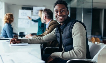 young businessman sitting in meeting with colleagues presenting in background. black man sitting in board room.