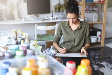 indoor view of beautiful young caucasian woman artist with brunette hair busy making drawings in spacious workshop interior with lots of paint bottles on table and computer monitor in background