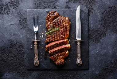 sliced grilled meat barbecue steak striploin with knife and fork carving set on black stone slate