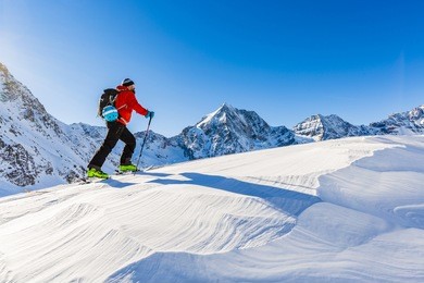 mountaineer backcountry ski walking up along a snowy ridge with skis in the backpack. in background blue sky and shiny sun and zebru, ortler in south tirol, italy.  adventure winter extreme sport. 