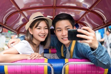 young asian couple tourists taking selfie while traveling on local colorful tuk tuk taxi exploring bangkok city, thailand