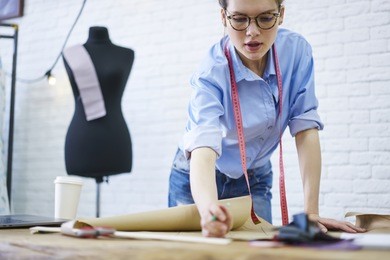 cropped image of skilled young woman in stylish eyeglasses and tape for measuring working dressmaker in fashion atelier standing at desktop and drawing sketch for collection of textile clothing 