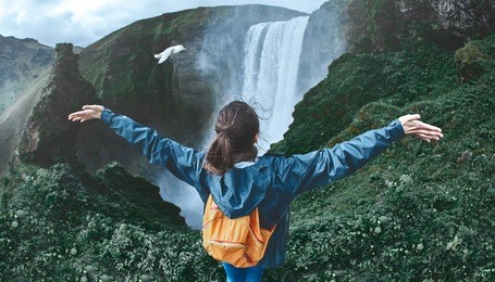 girl in waterproof clothing sits on the cliff on background of skogafoss waterfall in iceland. view from above