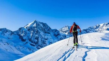 mountaineer backcountry ski walking up along a snowy ridge with skis in the backpack. in background blue sky and shiny sun and zebru, ortler in south tirol, italy.  adventure winter extreme sport. 