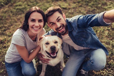 beautiful romantic couple is having fun with their dog labrador retriever outdoors. sitting on a green grass and making selfie.