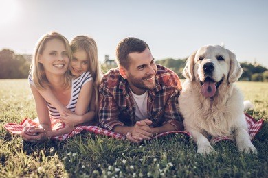 beautiful happy family is having fun with golden retriever outdoors. mother, father and daughter are lying with dog labrador on green grass in park.