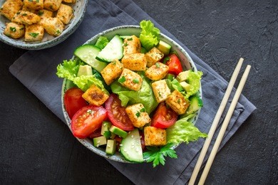 fried tofu salad with cucumbers, tomatoes, avocado and sesame seeds. homemade asian vegetable and tofu salad in ceramic bowl on black stone background. healthy asian diet vegan vegetarian salad food.