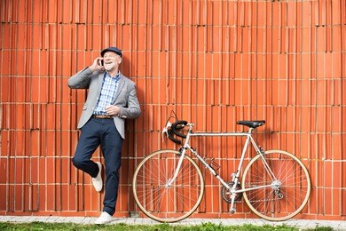 senior man with smartphone and bicycle against brick wall.