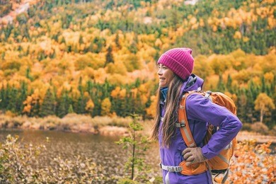 fall autumn hiker girl outdoor at nature forest lake with backpack for camping travel trip. happy asian woman hiking outdoors with bag and hat, cold outerwear gear.