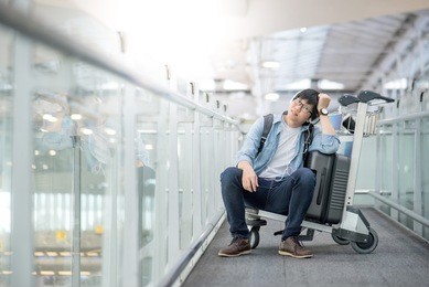 young asian man sitting on airport trolley with his suitcase luggage in the international airport terminal, waiting for travel abroad