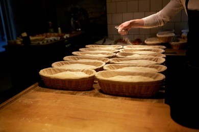 food cooking, baking and people concept - baker making bread and pouring flour to baskets while dough rising at bakery kitchen