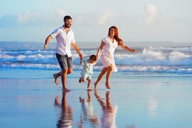 happy family - father, mother, baby son have fun together, child run with splashes by water pool along sunset sea surf on black sand beach. travel lifestyle, parents with kids on summer vacation.
