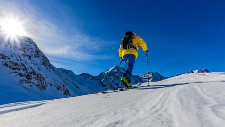 mountaineer backcountry ski spring walking up along a snowy ridge with skis in the backpack. in background blue cloudy sky and shiny sun and tre cime, drei zinnen in south tirol, dolomites, italy. 