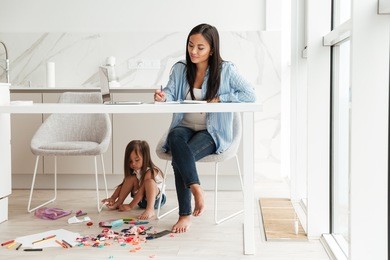 young pretty asian woman working with laptop and notepad while her little daughter playing on a floor at the kitchen