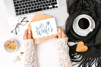 woman holding card with word hello winter. workspace with laptop,  coffee cup wrapped in scarf, and glasses. 