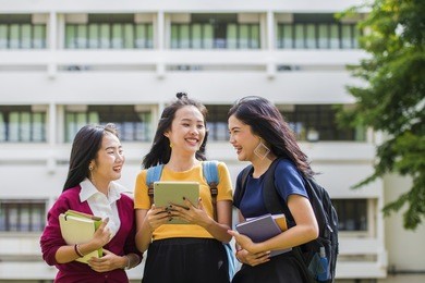 advertising, education students people knowledge concept - group of beautiful asian students talking together and checking on line content on tablet with the university building in the background. 