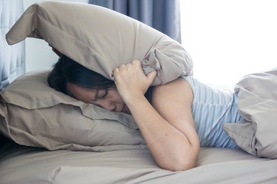 young woman lying in bed suffering from sound covering head and ears with pillow making unpleasant face. early wake up not getting enough sleep