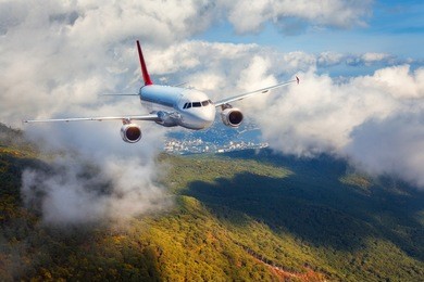 airplane is flying in clouds over mountains with forest at sunset. landscape with white passenger airplane, cloudy sky and green trees. passenger aircraft is landing. business travel. commercial plane