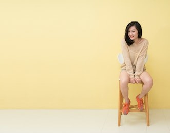 full length image of happy girl in long sleeve t-shirt and shorts on chair and looking at the camera over yellow background