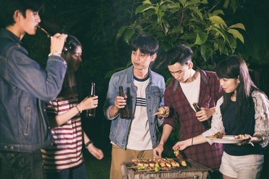 asian group of friends having outdoor garden barbecue laughing with alcoholic beer drinks on night 