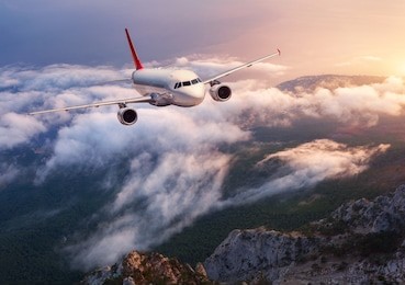 beautiful airplane is flying over low clouds at sunset. landscape with passenger airplane, rocks, forest, blue sky with orange sunlight in dusk. passenger aircraft. business travel. commercial plane