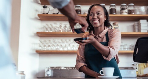 smiling barista using nfs technology to help a customer pay for a purchase with their bank card in a cafe 