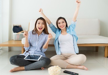 two girl friends sitting on the floor near the couch watch a movie and eat popcorn, relaxing together.