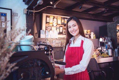 beautiful asian barista wearing red color apron working with coffee maker machine smiling at camera , small business concept.