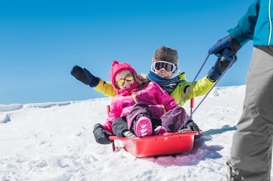 father sledding his little son and daughter. cute girl and happy boy being pulled on a red sled. dad pulling his children in a bobsled on the snow. winter vacation with family on snowy mountains.