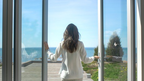 shot of a beautiful brunette with loose hair opening doors of the balcony and walking onto sunny terrace with seaside view. it's cloudless morning and she's really happy.