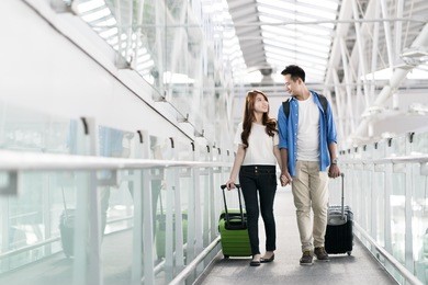 young happy asian couple carrying suitcase luggage in airport terminal. couple holding hand and traveling abroad together, air travel or holiday vacation concept